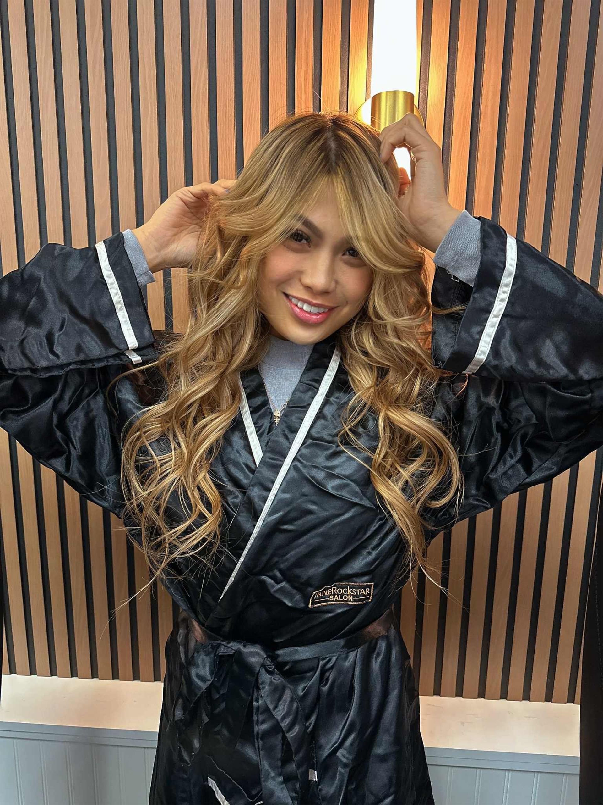 Woman in black robe with blonde curly hair smiles, hands near head, against a striped backdrop.