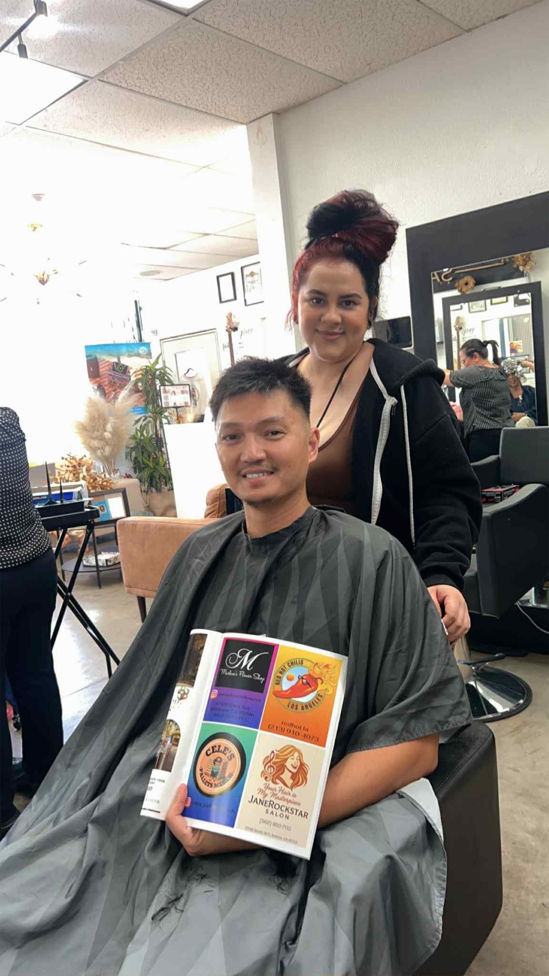 Man getting a haircut at a salon, holding a magazine, with hairstylist smiling behind him.