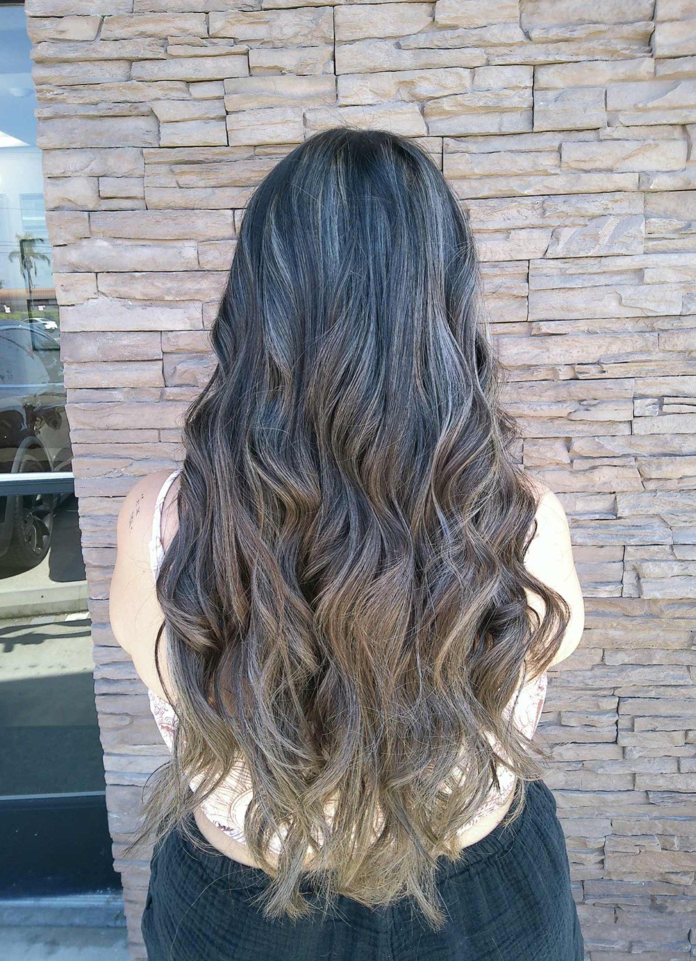 Woman with long wavy hair in brown ombre style, standing in front of a stone wall.