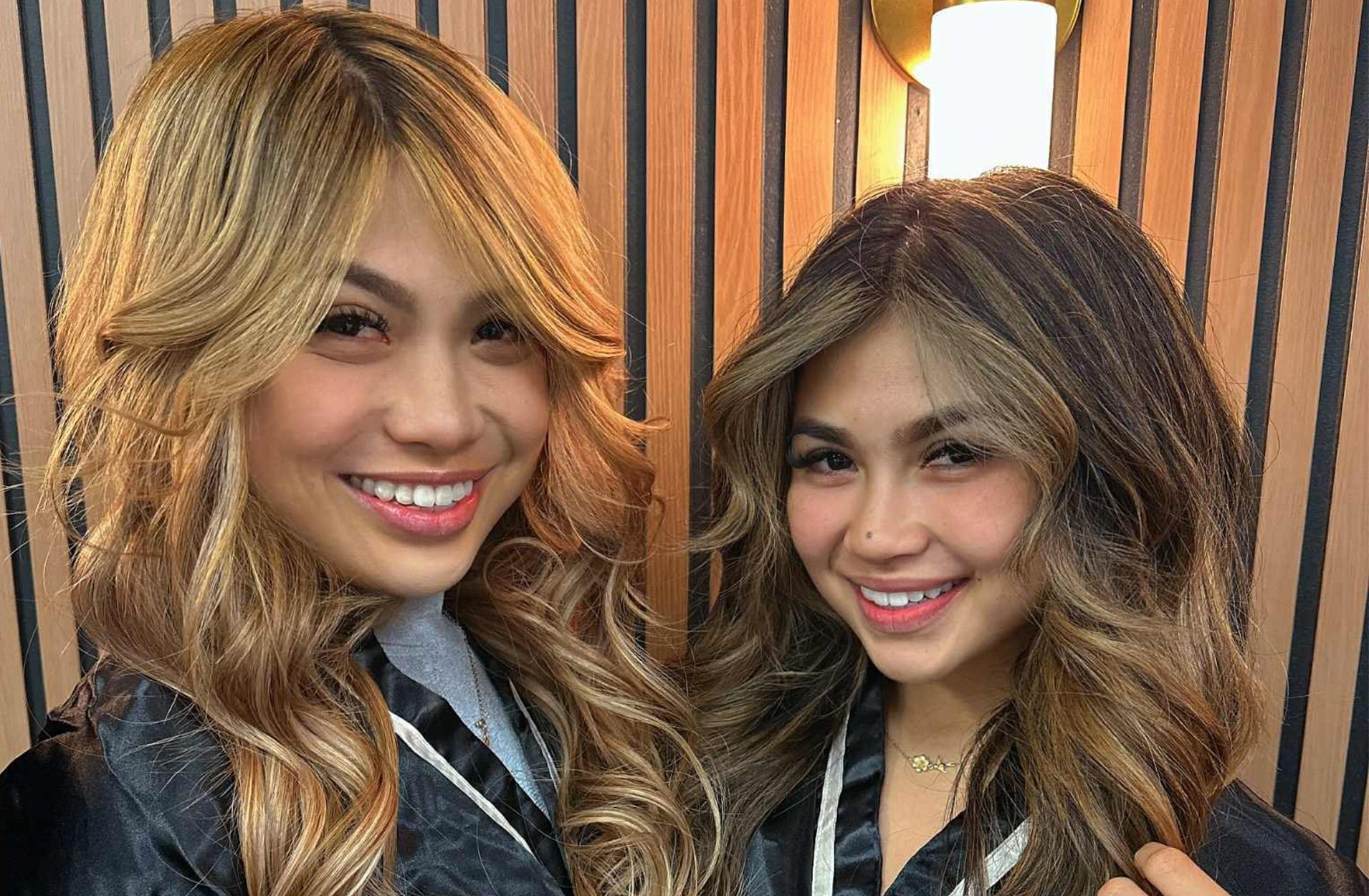 Two smiling women with new highlighted hairstyles in a salon, near a wooden wall.