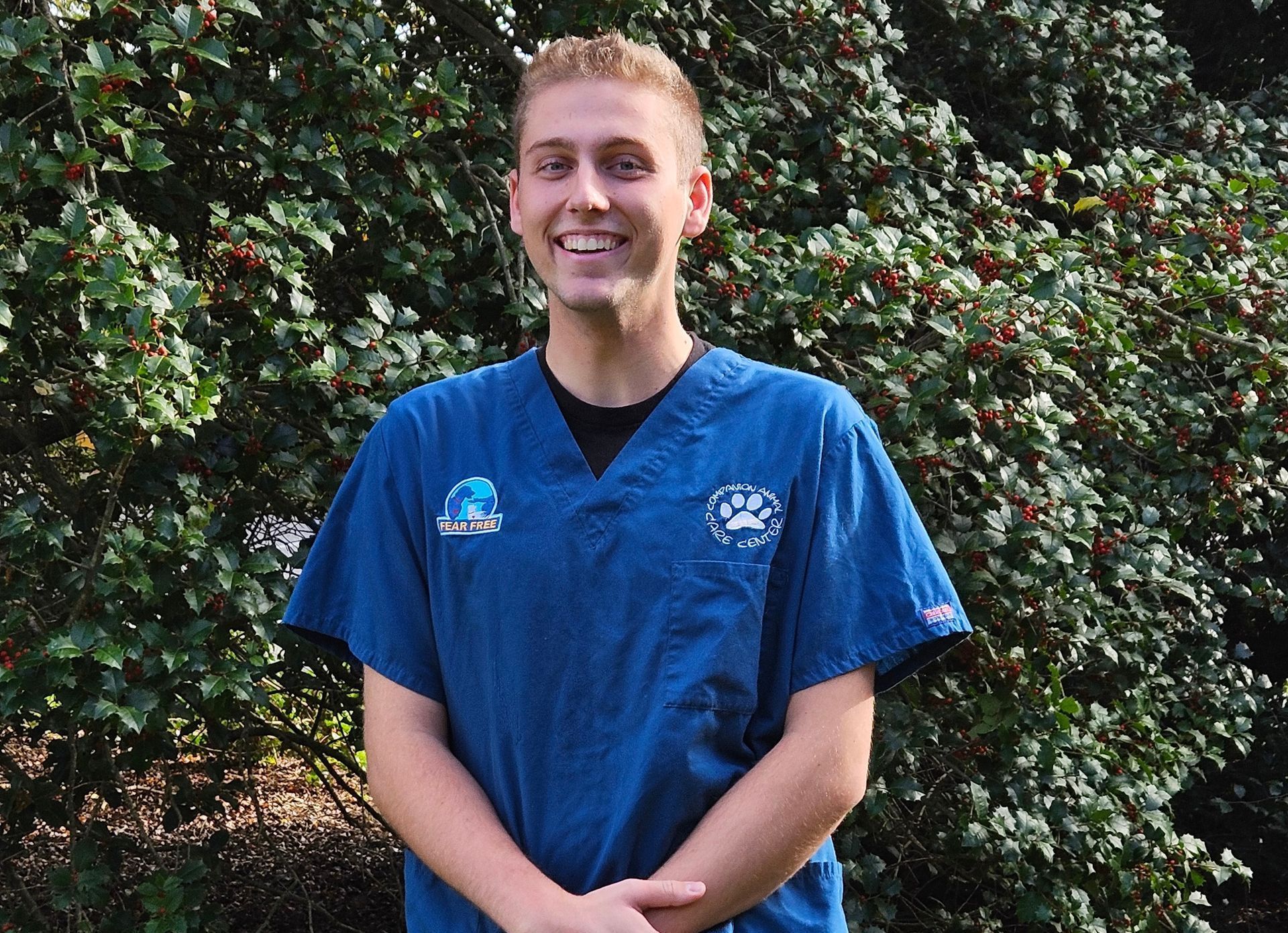 A woman in a blue scrub top is standing in front of a bush.
