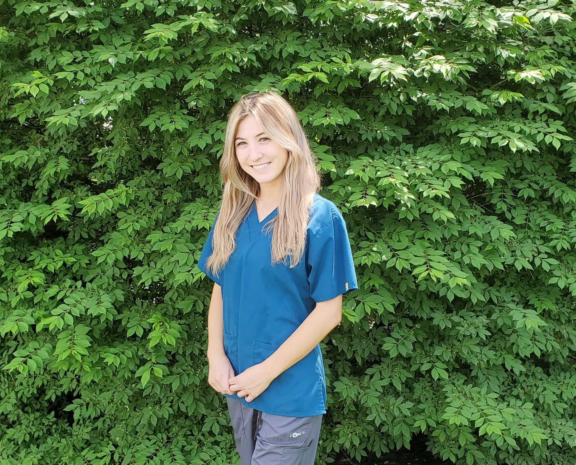 A young woman in a blue scrub top is standing in front of a tree.