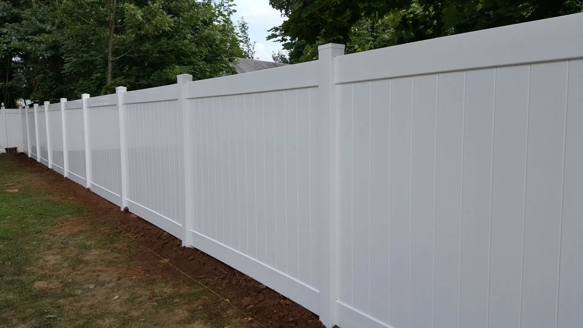 A white vinyl fence surrounds a backyard with trees in the background.