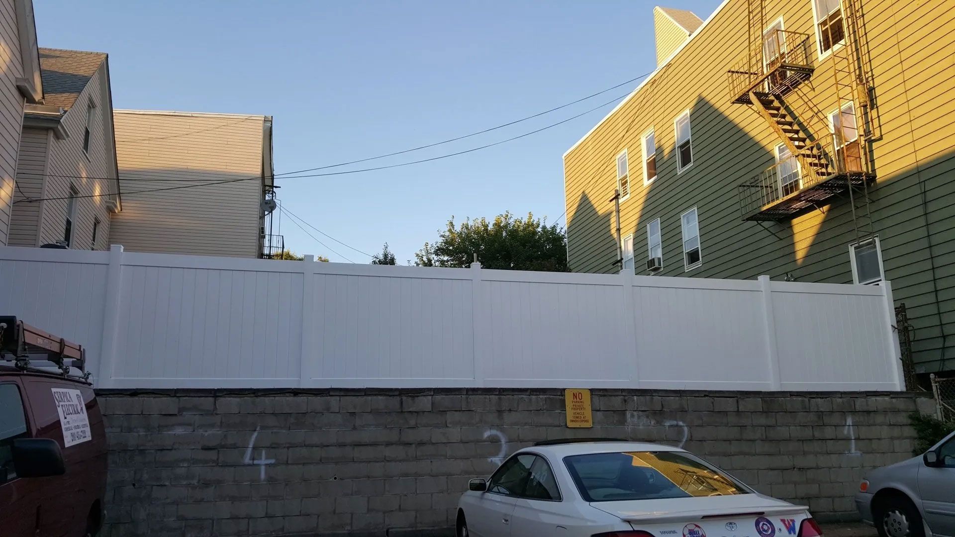 A white car is parked in front of a white fence.