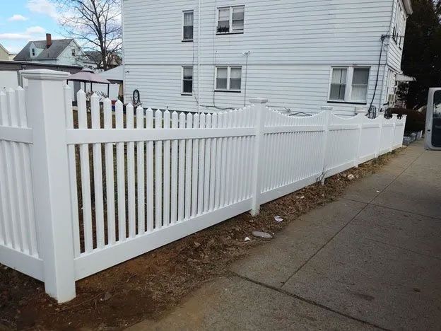 A white picket fence surrounds a white house on a sidewalk.