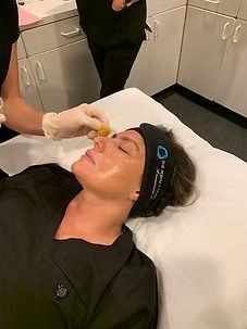 Person lies on a treatment bed while another applies a facial treatment in a clinic.