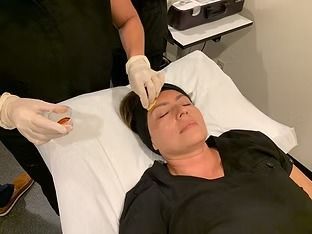 Person receiving a facial treatment while lying on a treatment table, with gloved hands applying product to the forehead.