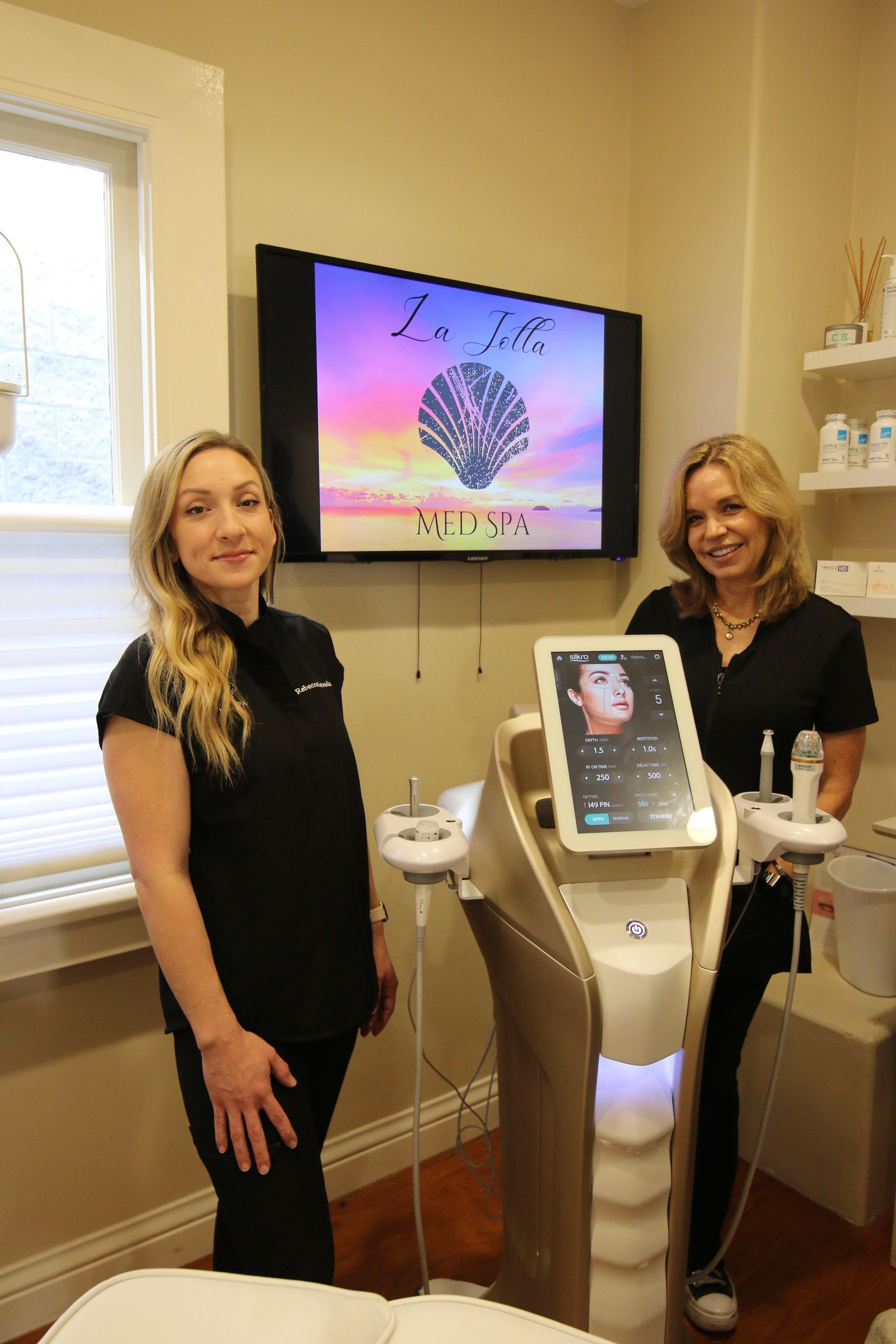 Two women standing beside a facial treatment machine in a spa room, with a wall-mounted screen behind them.