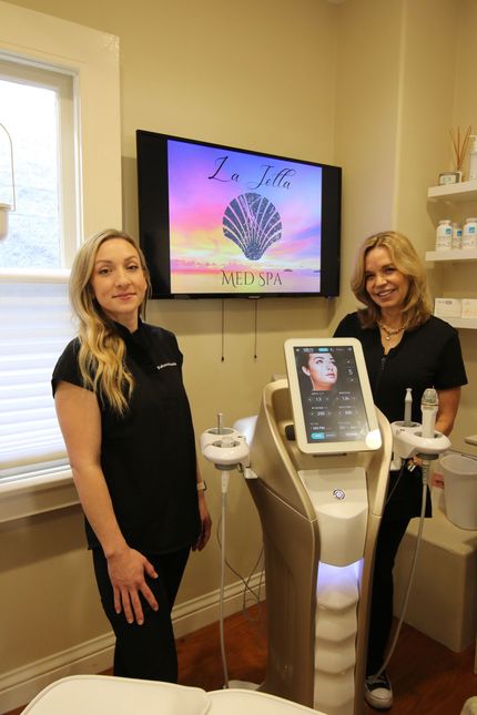 Two women standing beside a facial treatment machine in a spa room, with a wall-mounted screen behind them.