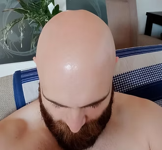 Person with a bald head and beard looking down, seated indoors on a bed or couch