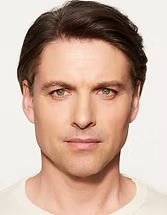 Close-up portrait of a person with short dark hair and a neutral expression on a plain light background