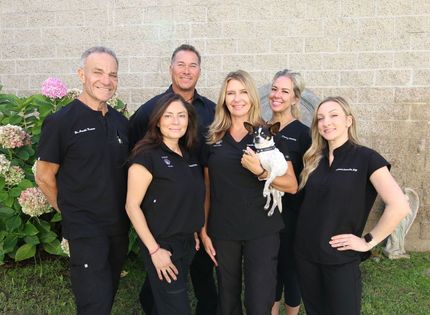 Six people in black uniforms posing outdoors, one holding a small dog, against a stone wall