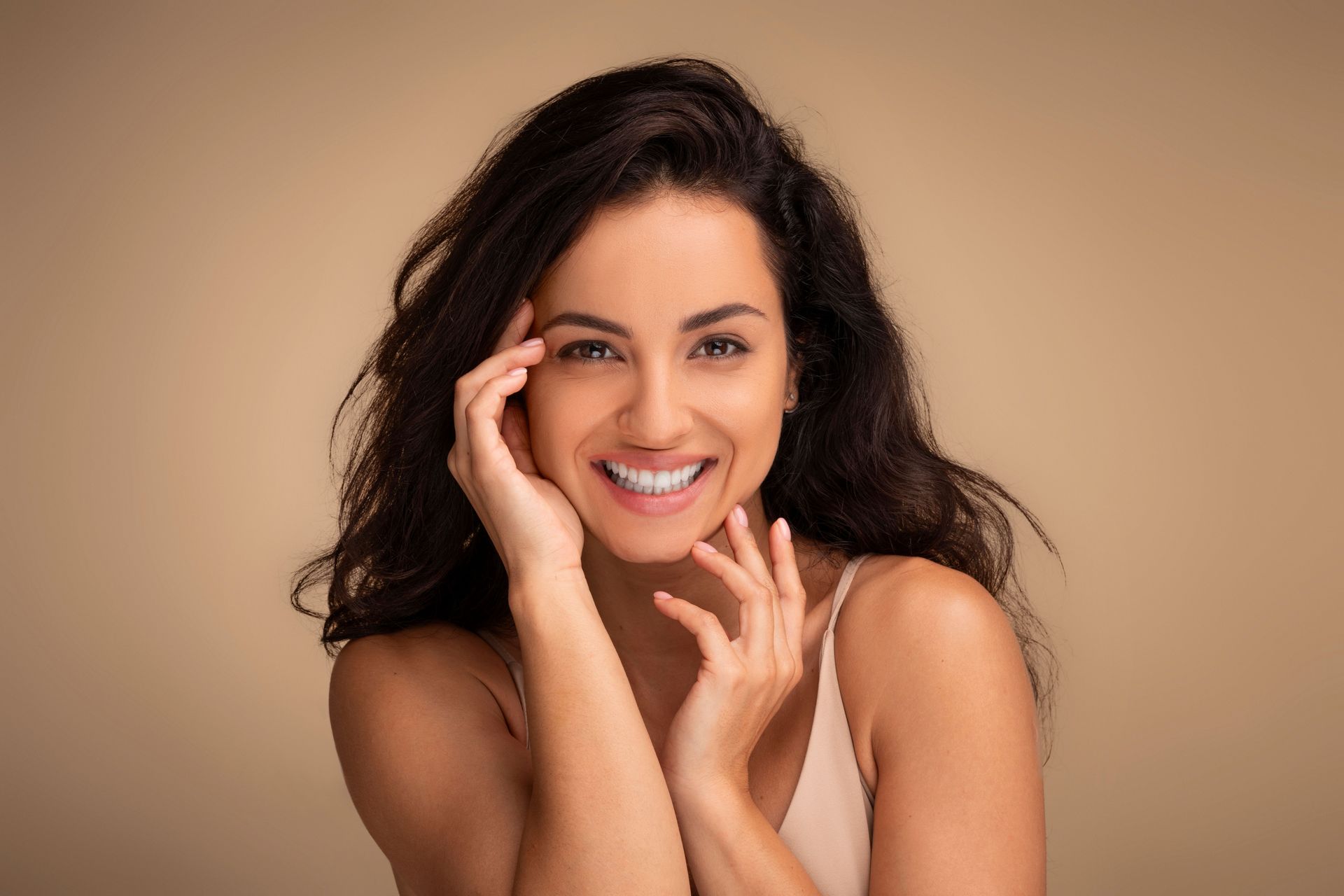 Smiling woman with dark hair posing against a beige background, hand on her face.