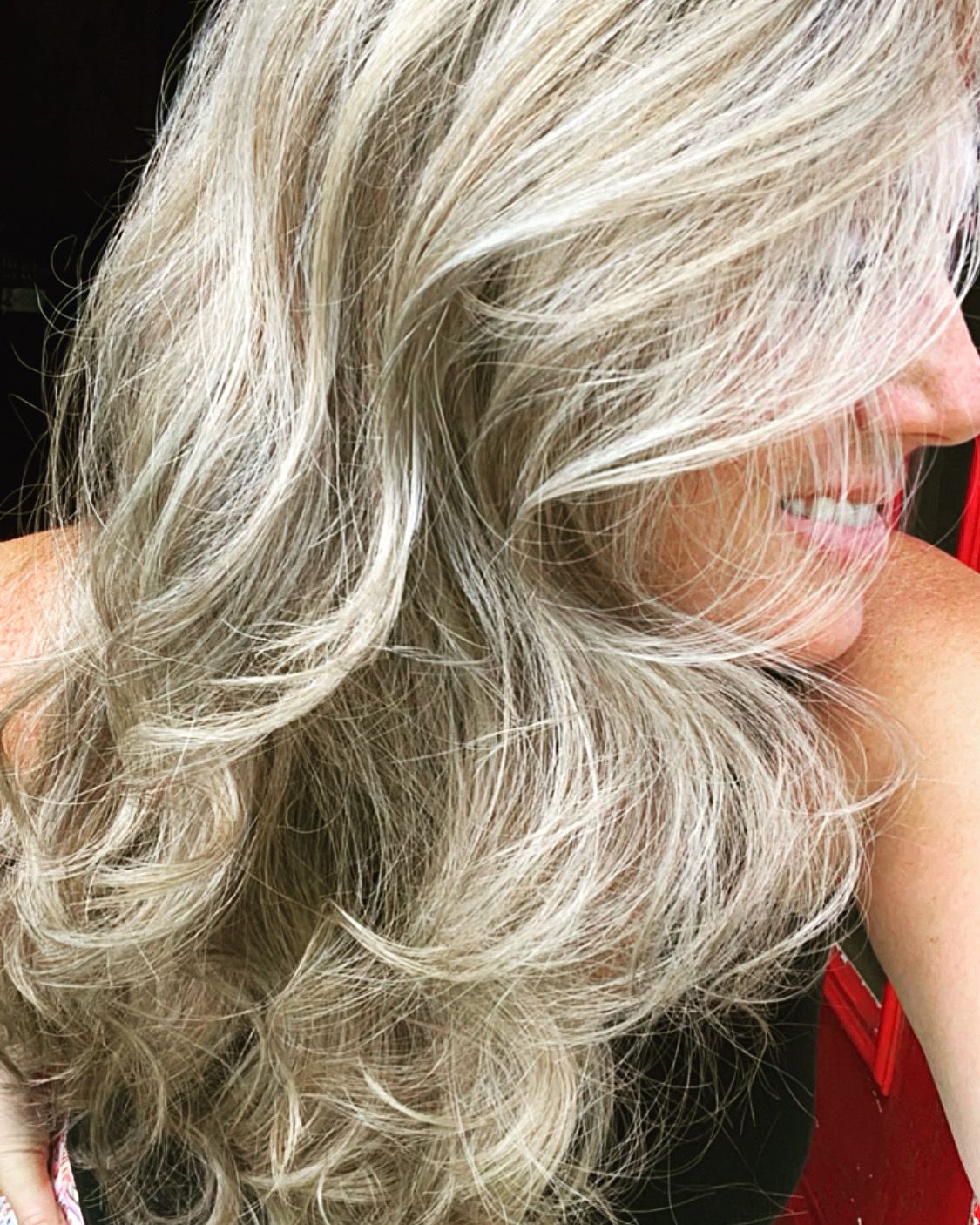 A woman with long blonde hair is smiling and holding her hair.