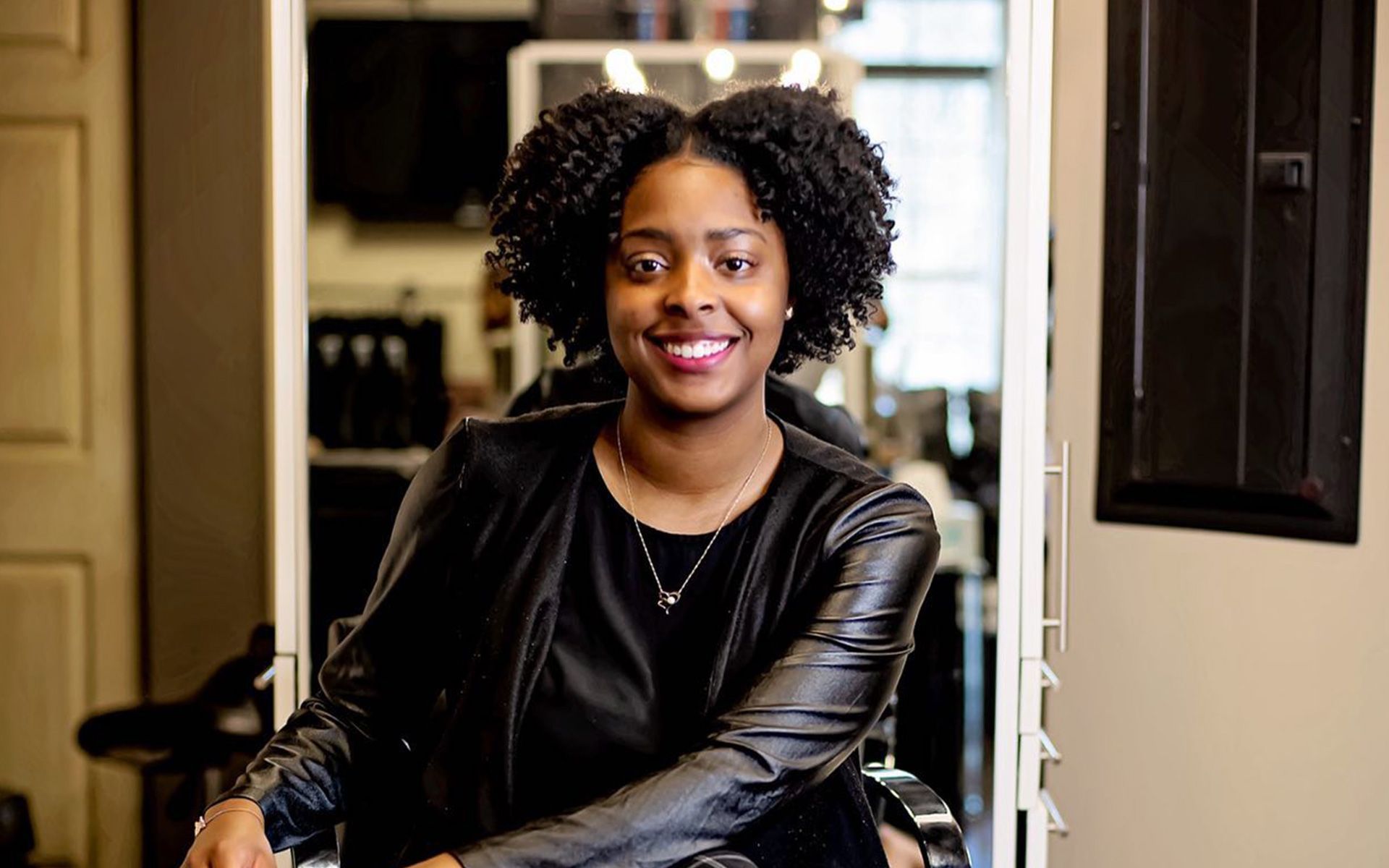 A woman in a black leather jacket is sitting in a chair in a salon.