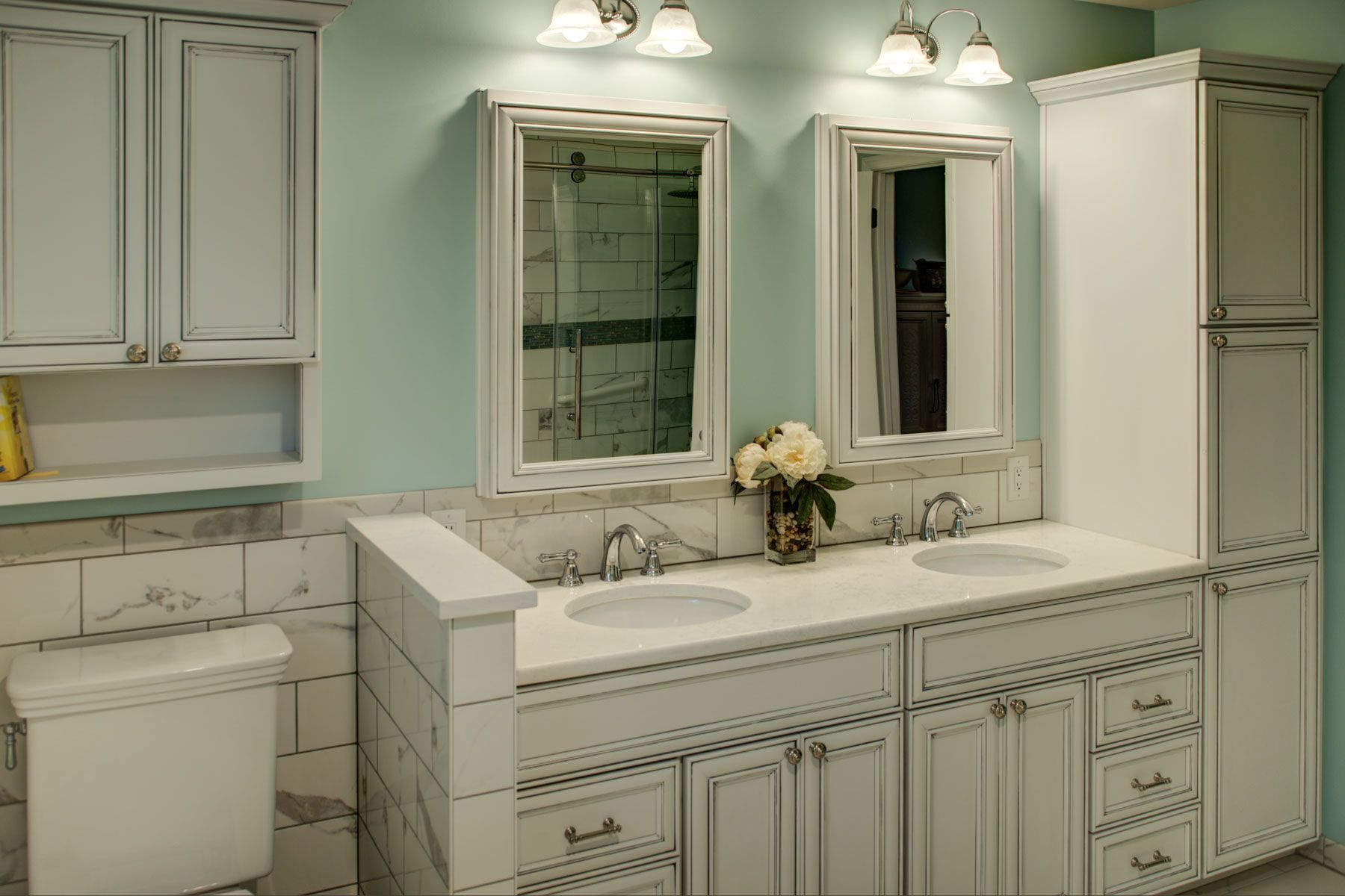 Bathroom with white cabinets, light blue wall, marble counter, two sinks, and mirrors
