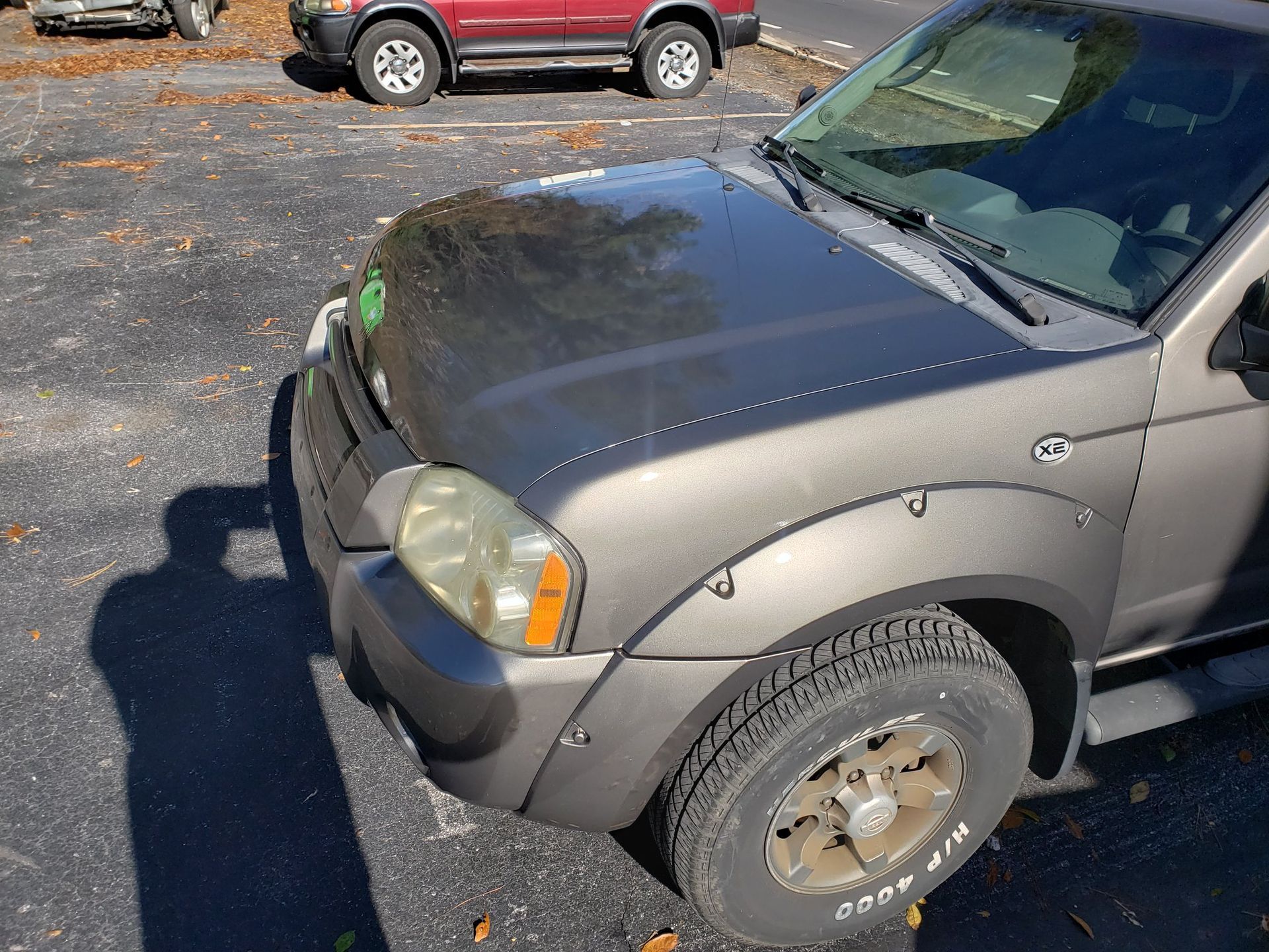 Gray Nissan Frontier truck parked in a lot; another red vehicle in the background. Sunny day.