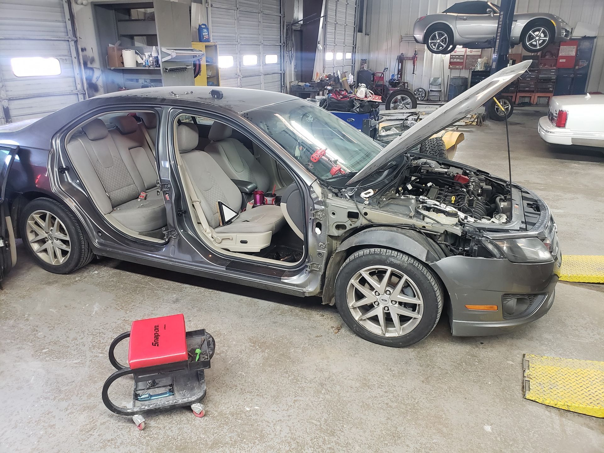 A gray car in a repair shop with doors and front quarter panels removed, hood open; a welding machine is in front.