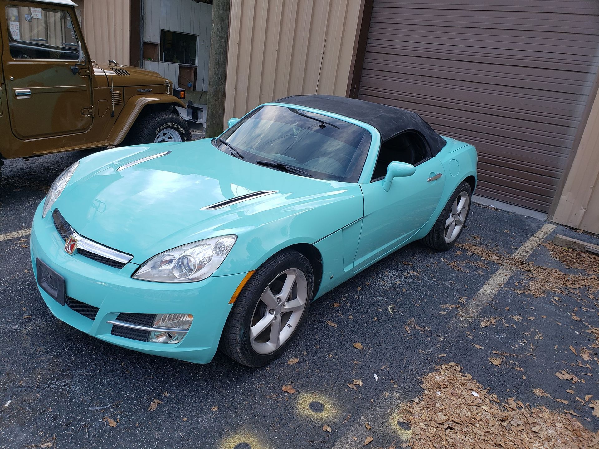 A turquoise Saturn Sky convertible parked next to a brown jeep, with a black roof, outside.