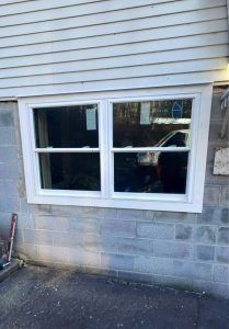 A white double-hung window installed in a light gray concrete block foundation wall beneath light-colored siding.