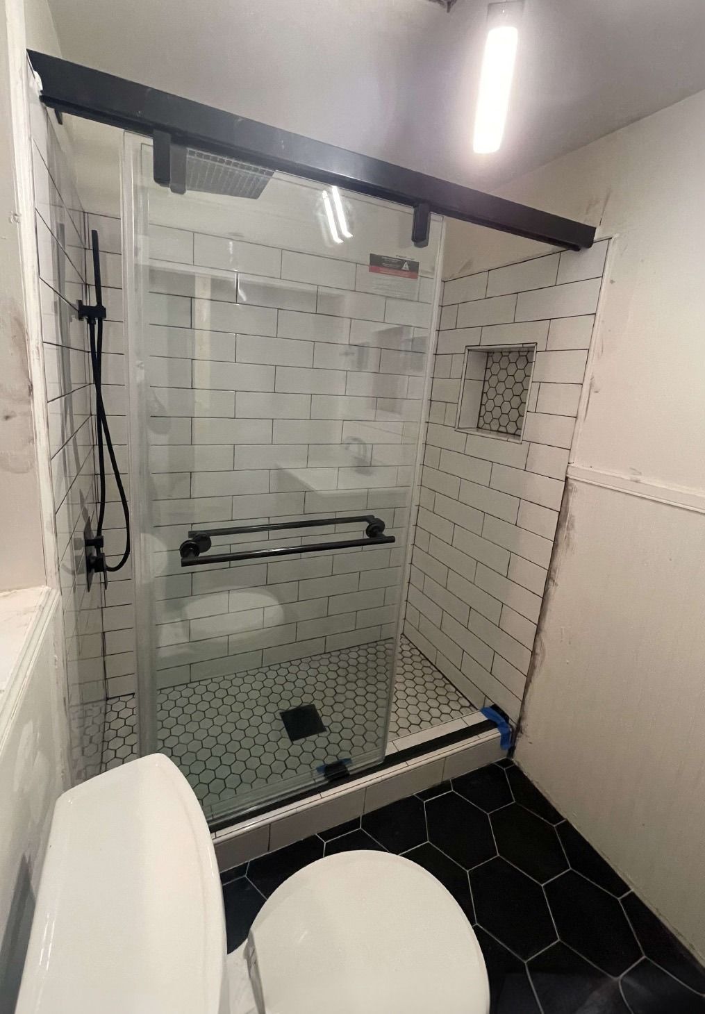 A walk-in shower with white subway tile, a black frame sliding glass door, and black hexagonal floor tiles.
