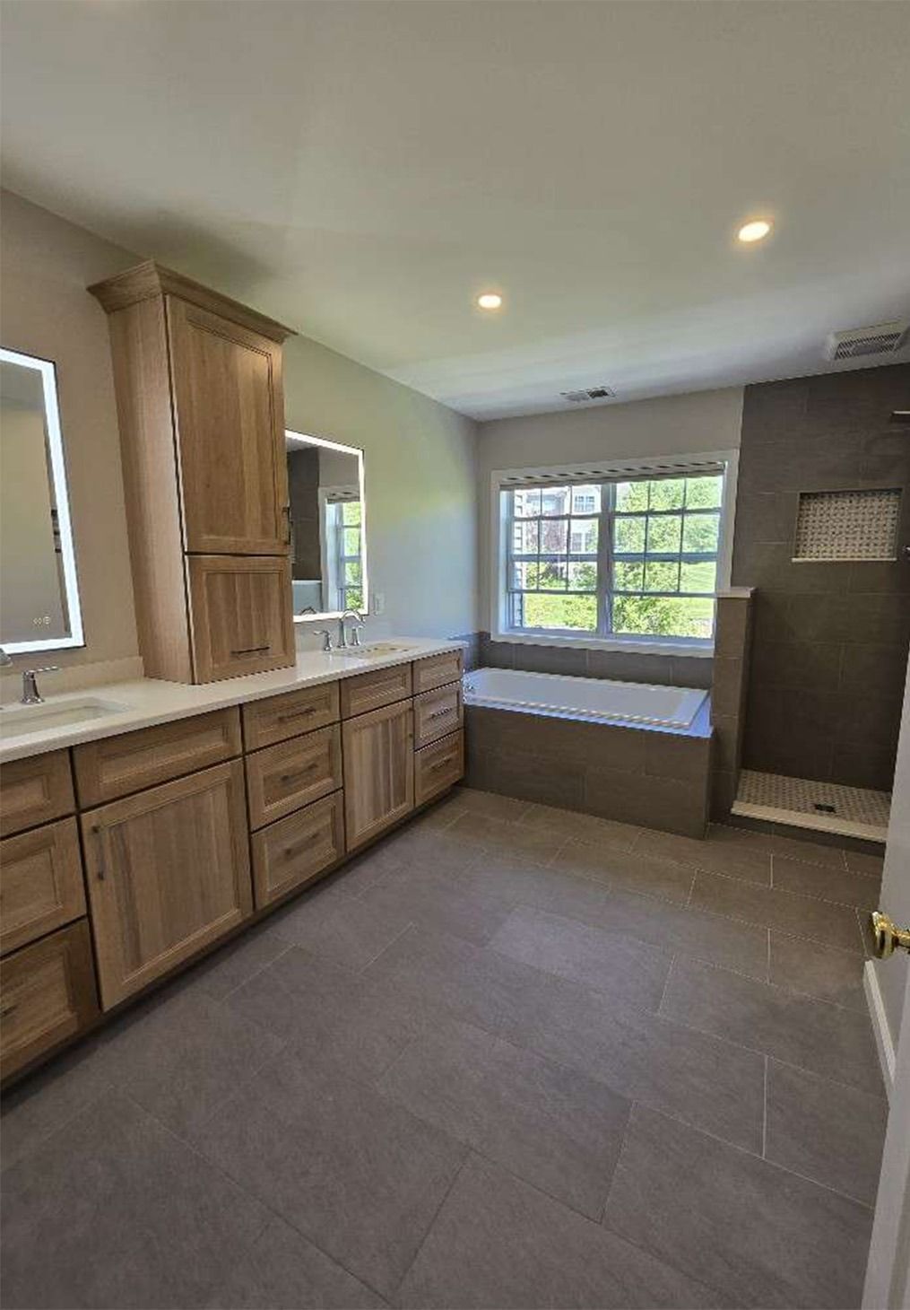 A spacious bathroom with wood cabinets, light countertops, two mirrors, a gray tiled floor, and a built-in bathtub.