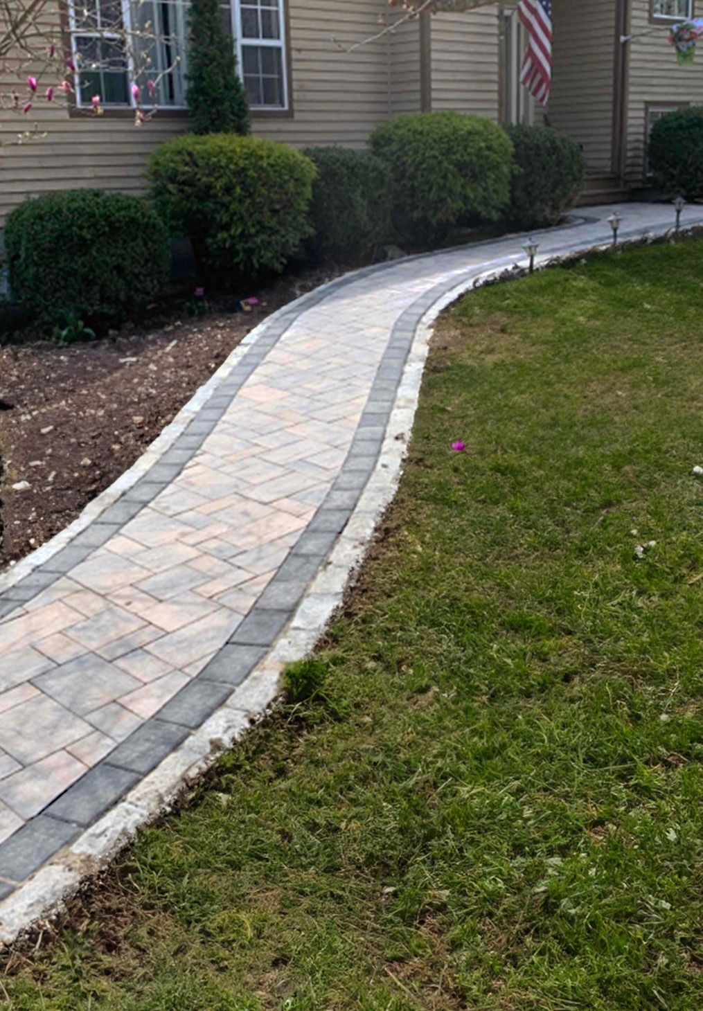 A curving paver walkway with a dark border leads toward the front entrance of a house lined with manicured bushes.