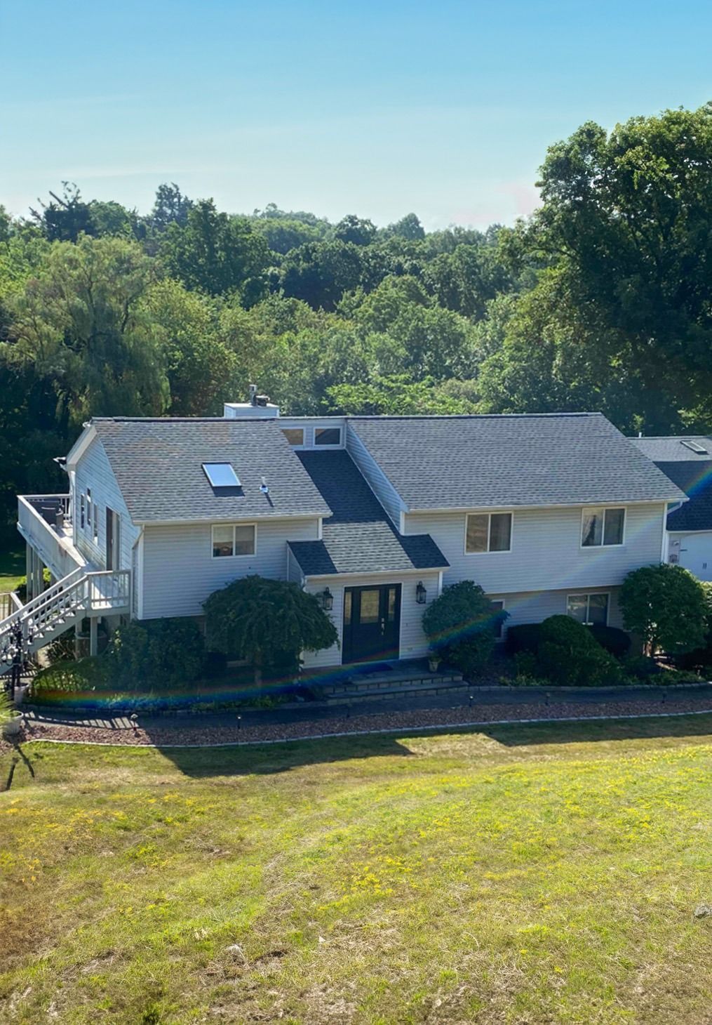 A light-gray multi-level house with a dark shingled roof, surrounded by trees under a clear blue sky.