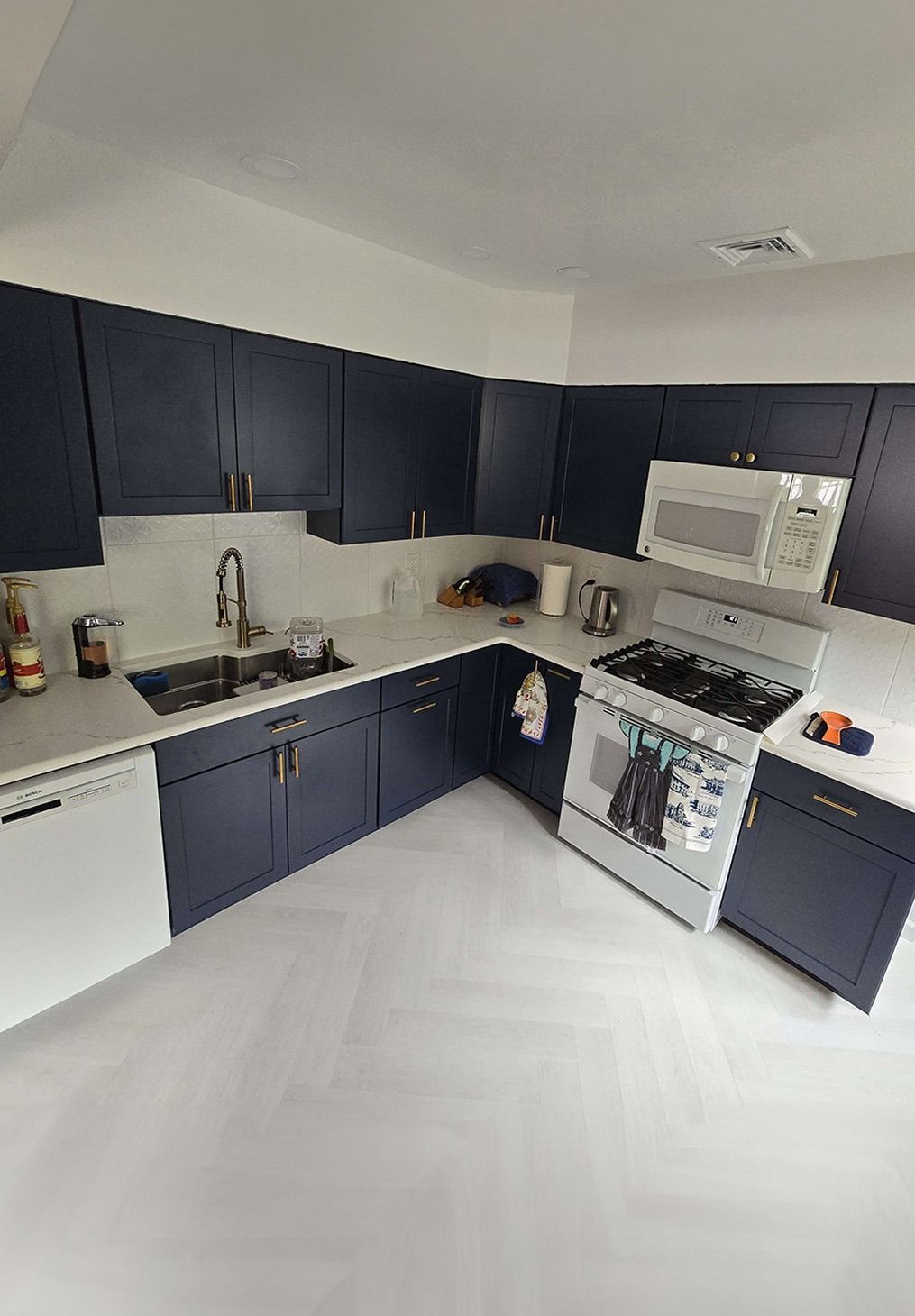 A kitchen with dark navy cabinets, white countertops, a white herringbone floor, and white appliances.