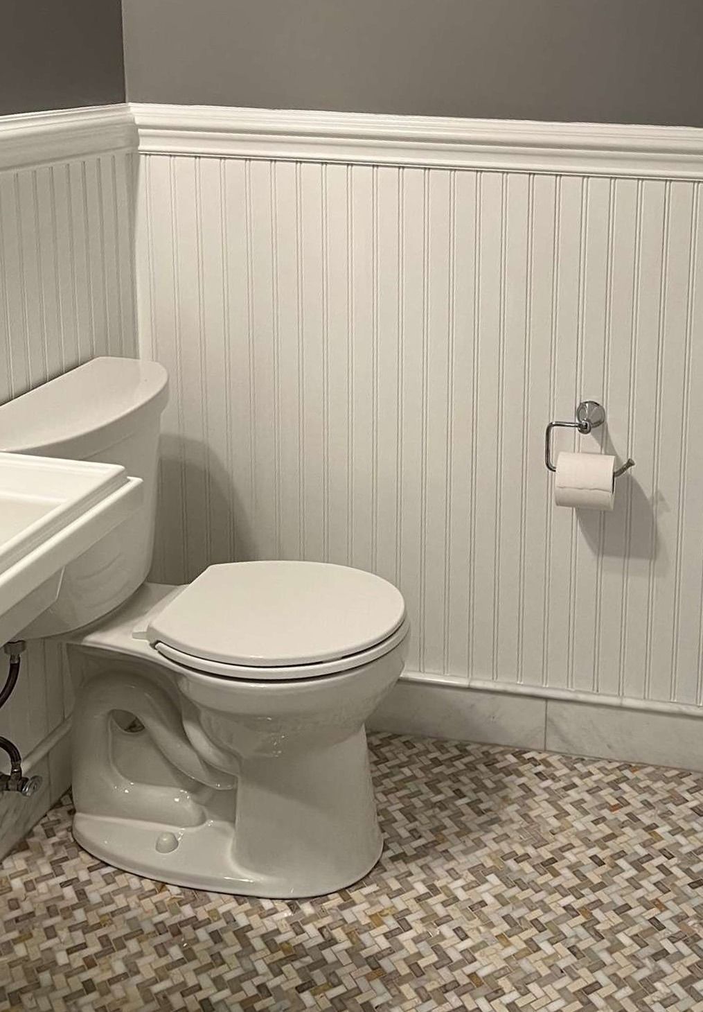 A toilet beside a wall with white vertical wainscoting and a toilet paper holder, above a patterned tile floor.