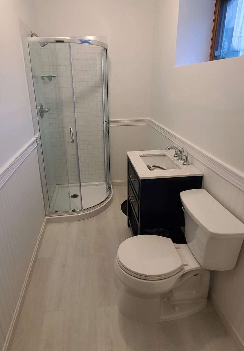 A bathroom with a corner shower, a dark vanity with a white countertop, and a white toilet on light wood-look flooring.
