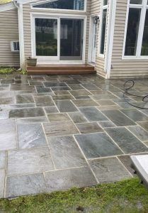 A light-colored house with a sliding glass door and a low wooden step leading onto a large, gray flagstone patio.