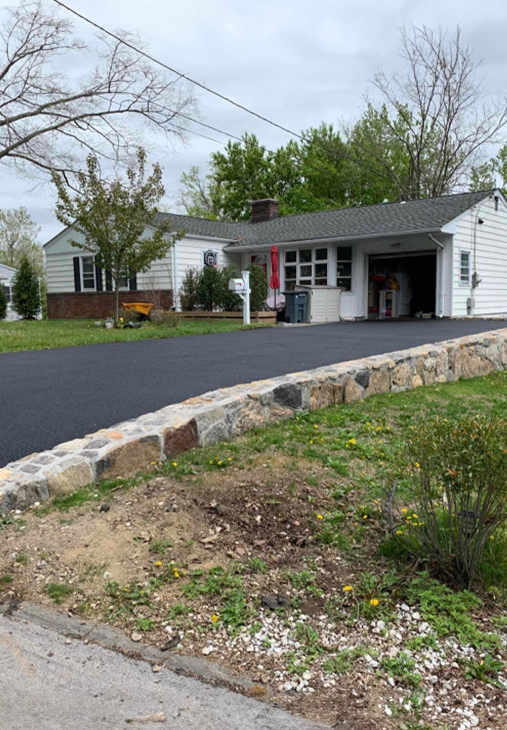 A single-story white house with a stone retaining wall along a fresh asphalt driveway and a built-in garage.
