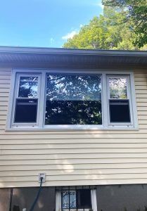 A three-pane window with light gray frames set against light yellow horizontal vinyl siding on an exterior wall.