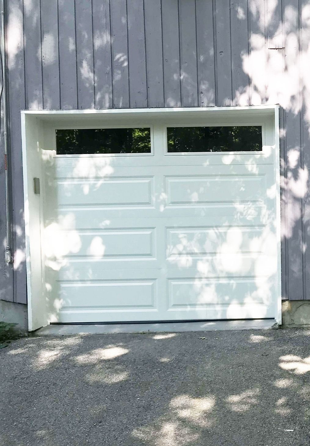 A white, two-panel garage door with rectangular windows at the top, set into a grey wooden-paneled building.