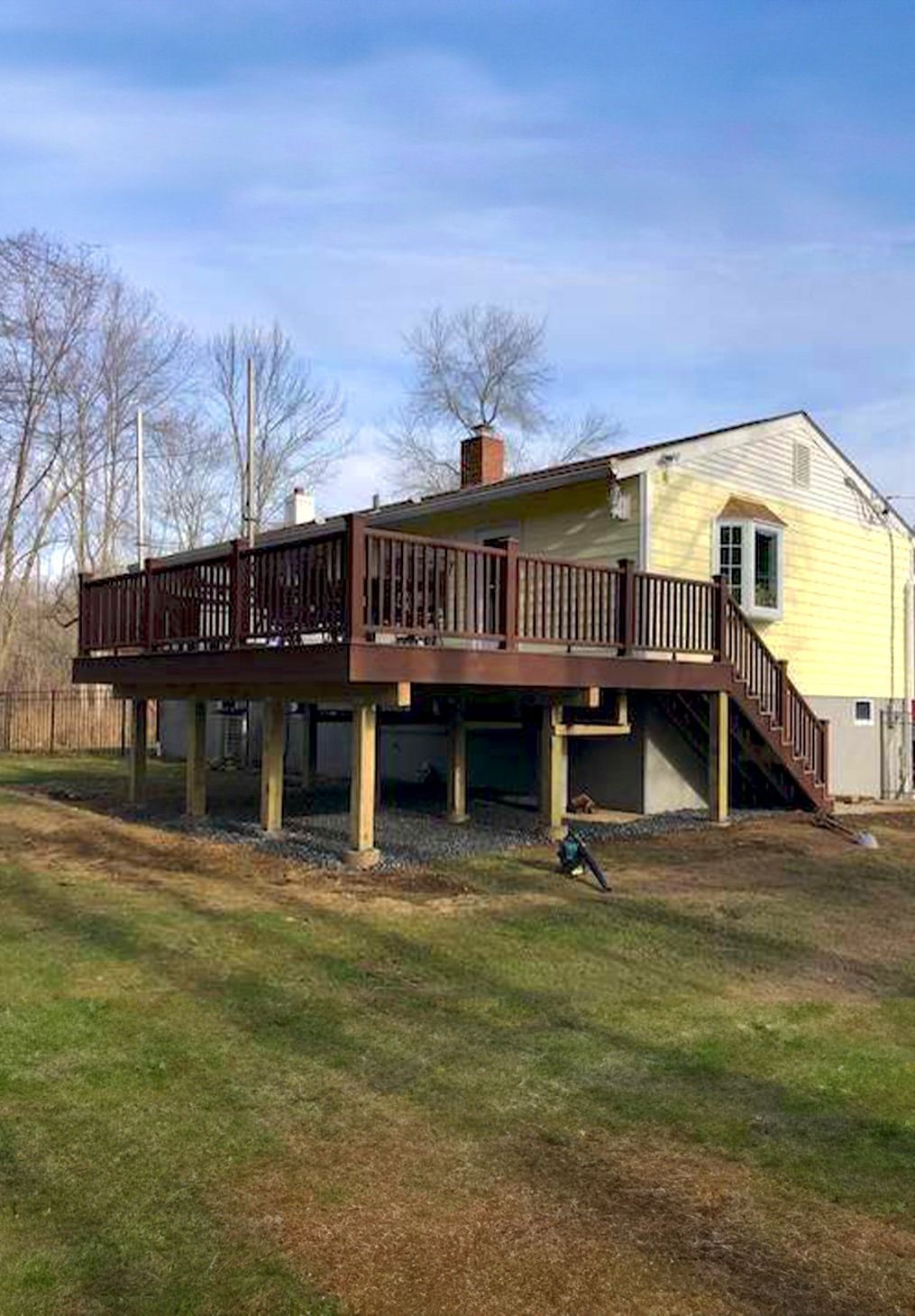 A light yellow house features a large, elevated dark brown wooden deck supported by posts, with stairs leading to the yard.