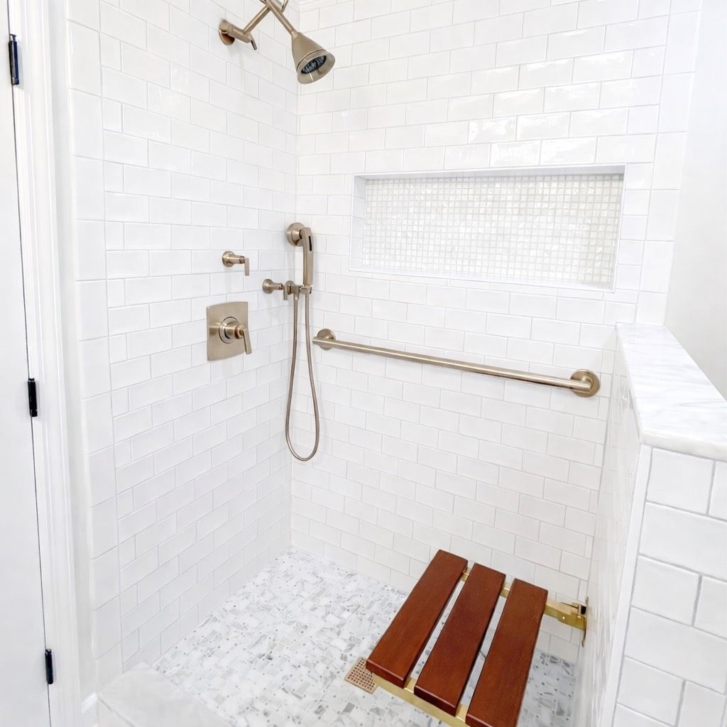 Walk-in shower with white subway tiles, a gold showerhead, handheld sprayer, grab bar, and a wooden folding bench.