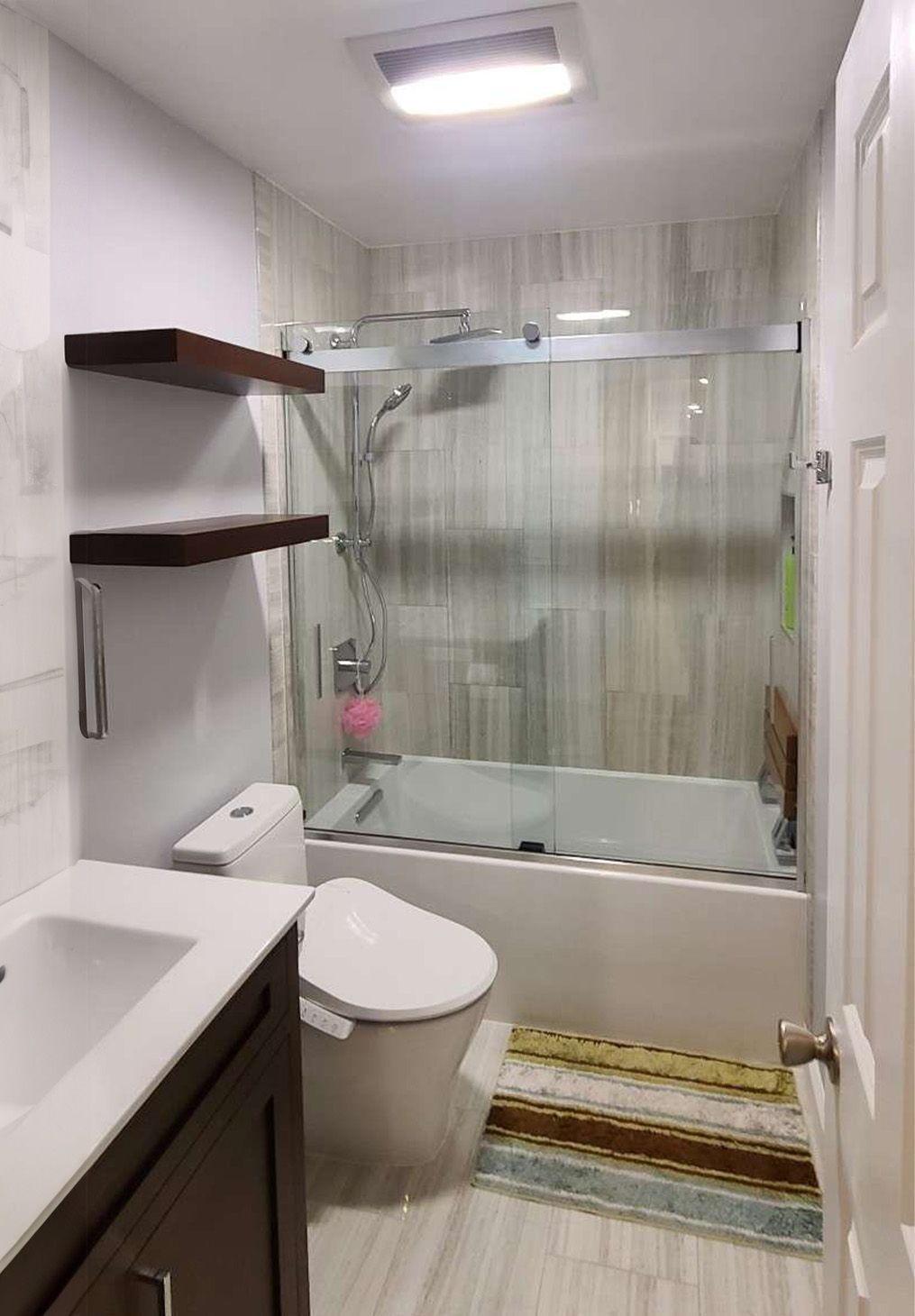 A small, modern bathroom featuring a white vanity, a toilet, and a glass-enclosed bathtub with a shower head.
