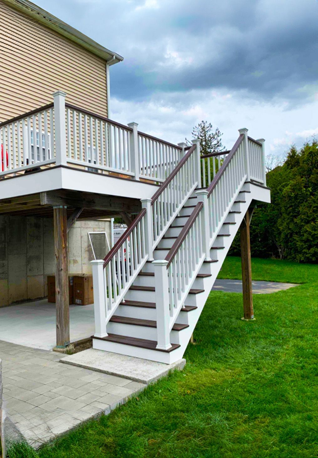 A raised wooden deck with white railings and brown treads, connected to a house via a matching staircase to a patio.