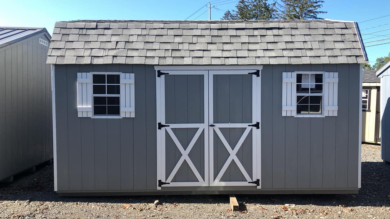 A gray shed with white trim and shutters is sitting on top of a gravel lot.