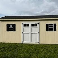 A white shed with black shutters is sitting in the middle of a grassy field.