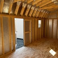 A gray shed with white doors and windows is sitting in the middle of a forest.