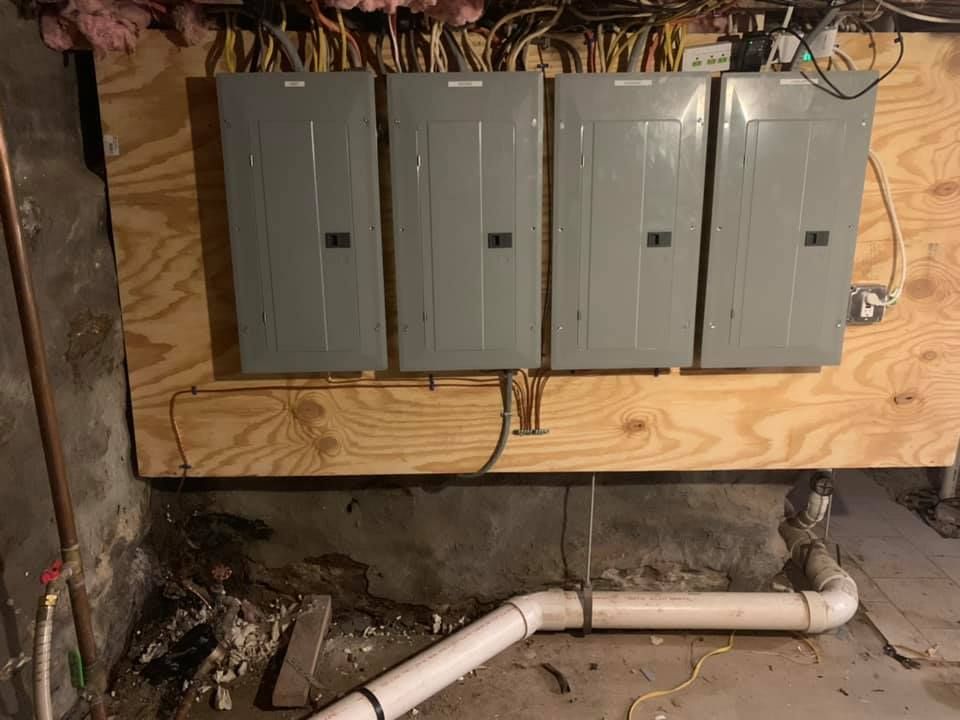 Three electrical panels are hanging on a wooden wall in a basement.