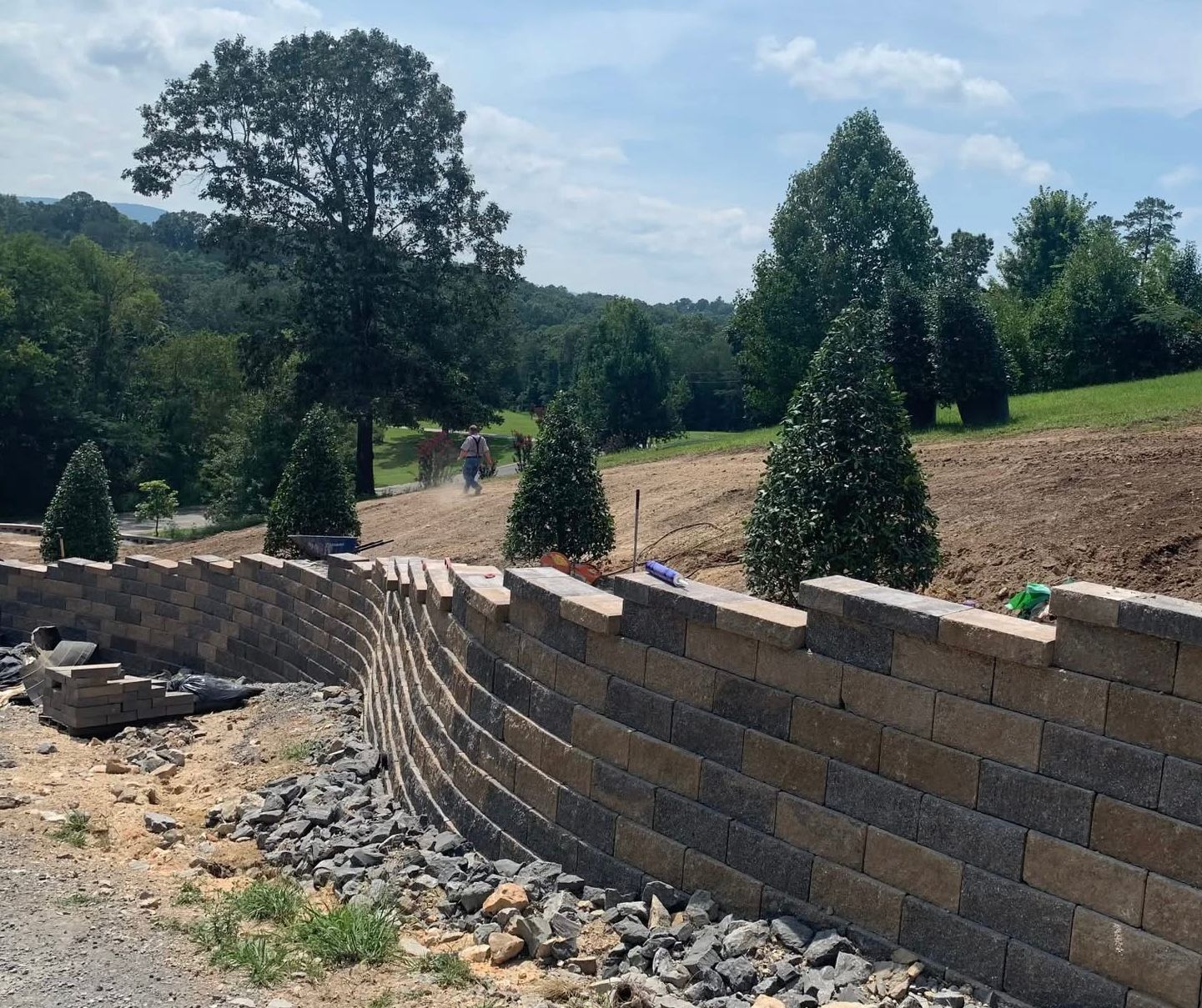 A brick wall is being built on a hillside with trees in the background.