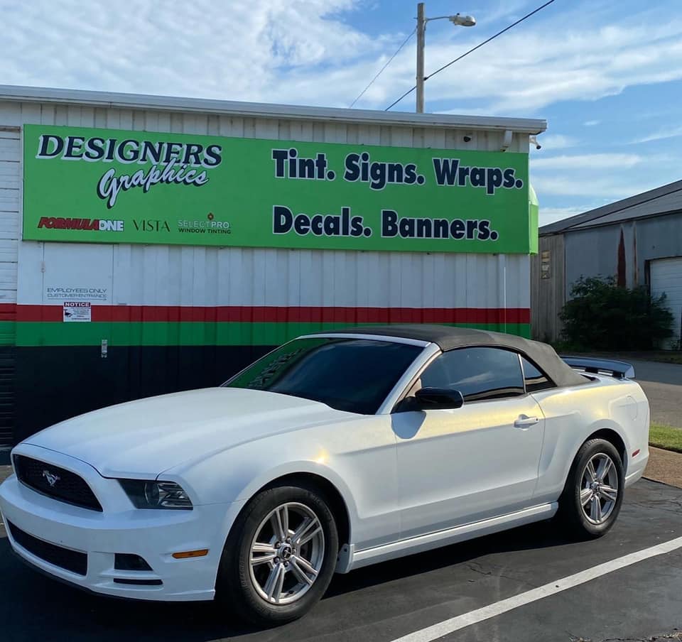 A white convertible mustang is parked in front of a sign that says designers tint signs wraps decals banners