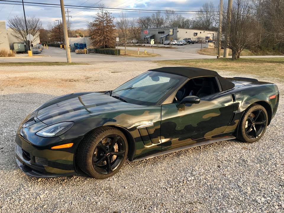 A camouflaged sports car is parked in a gravel lot.