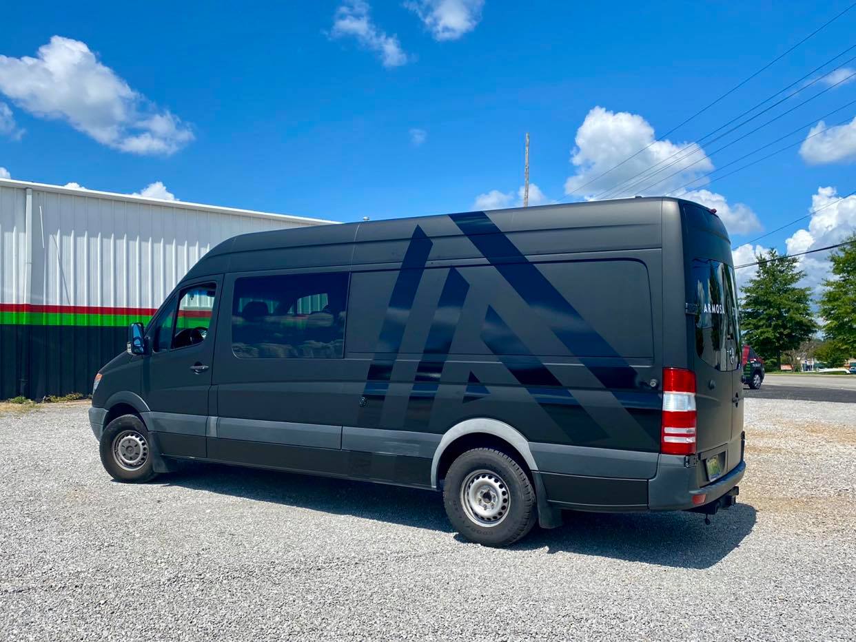 A black van is parked in a gravel lot in front of a building.