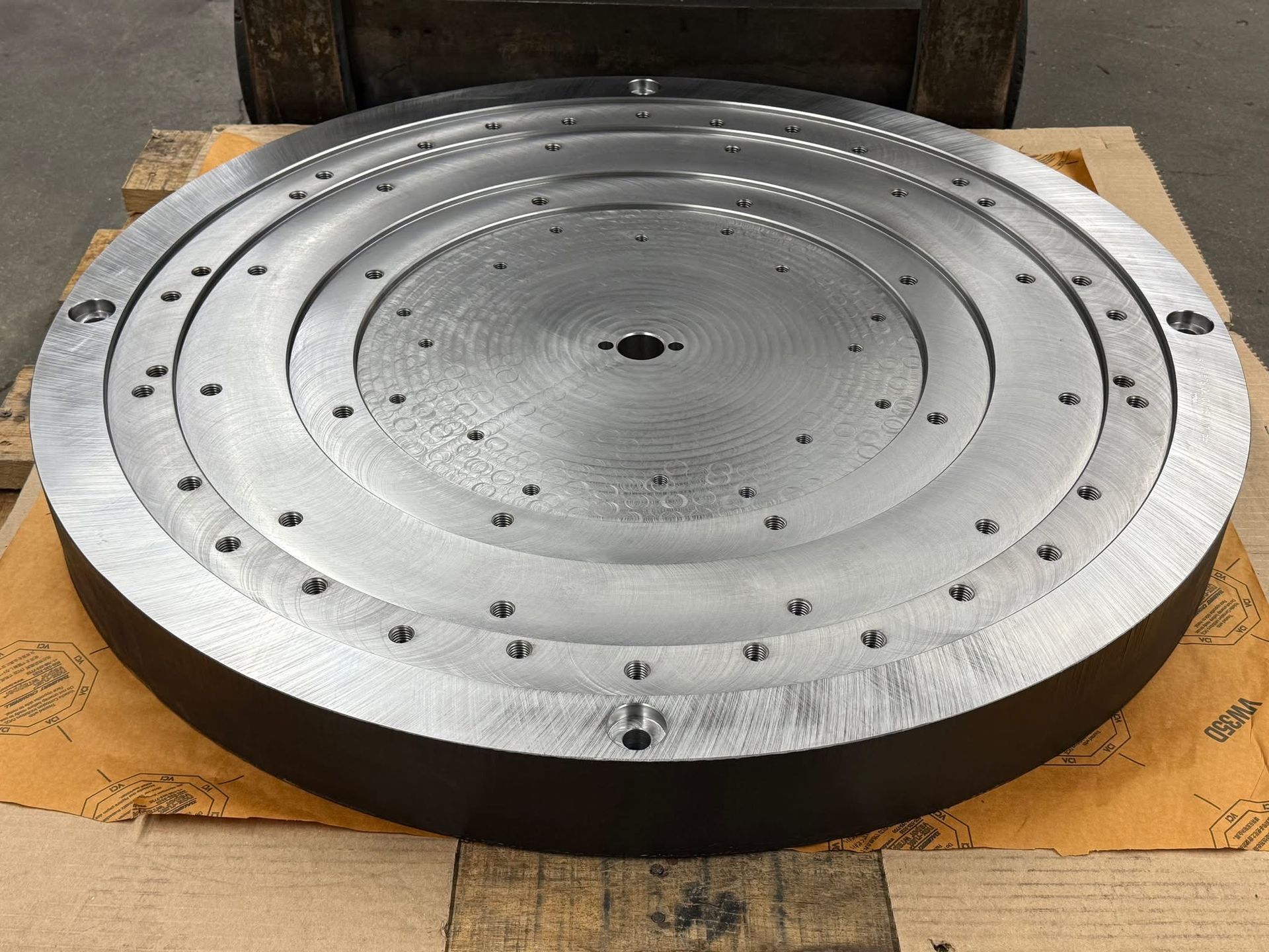 A large, circular, machined steel industrial component with bolt holes rests on a pallet.