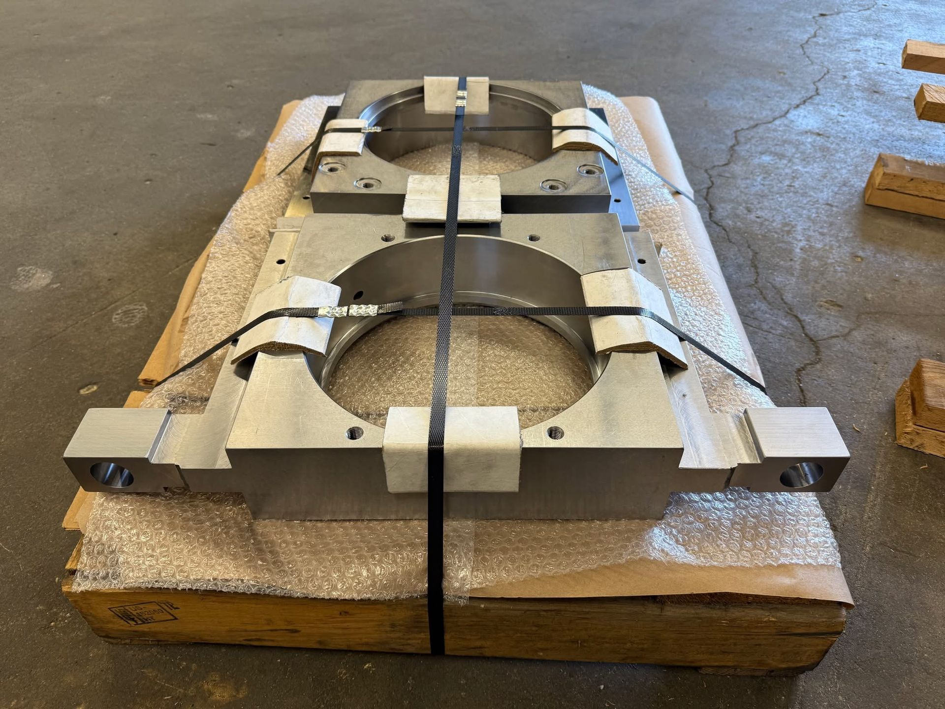 A heavy, machined metal part with two large circular openings, secured with black straps on a wooden pallet.