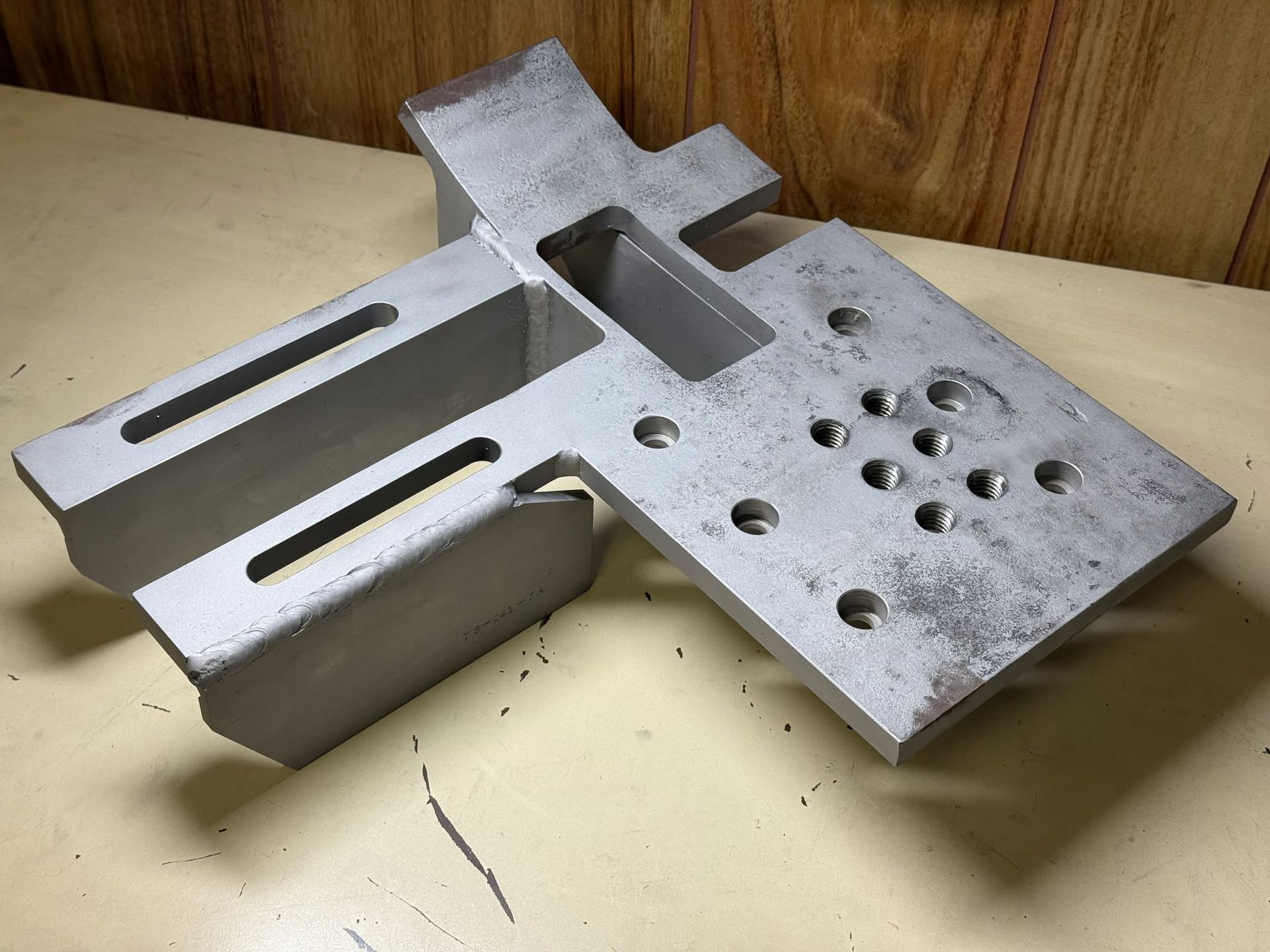 A metal mechanical mounting plate with slotted arms and a patterned array of drill holes, resting on a flat surface.